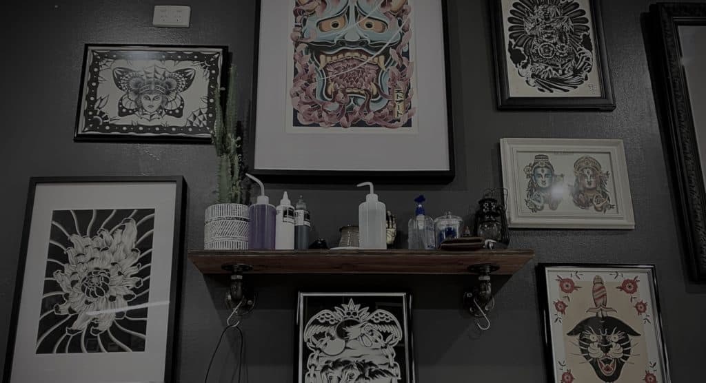 Framed tattoo art of various styles displayed on a wall, with tattoo cleaning products and inks sitting on a shelf. Taken at the Anchored Arts Tattoo Studio in Buddina, Sunshine Coast